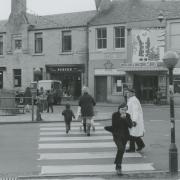 This photograph shows people crossing at a zebra crossing on High Street, Tranent. In the background are the shops of P. & H. Marr (flowers), Purves (drapery), George McNeill (plasterers), Tranent Cinema and J. Williams Footwear.