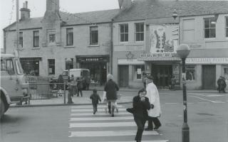 This photograph shows people crossing at a zebra crossing on High Street, Tranent. In the background are the shops of P. & H. Marr (flowers), Purves (drapery), George McNeill (plasterers), Tranent Cinema and J. Williams Footwear.